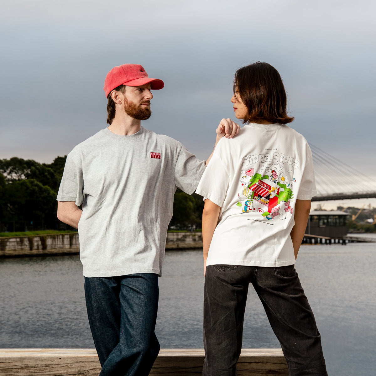 Two people standing on a wooden dock by a body of water, wearing white and grey Sippa Sippa Park t-shirts.