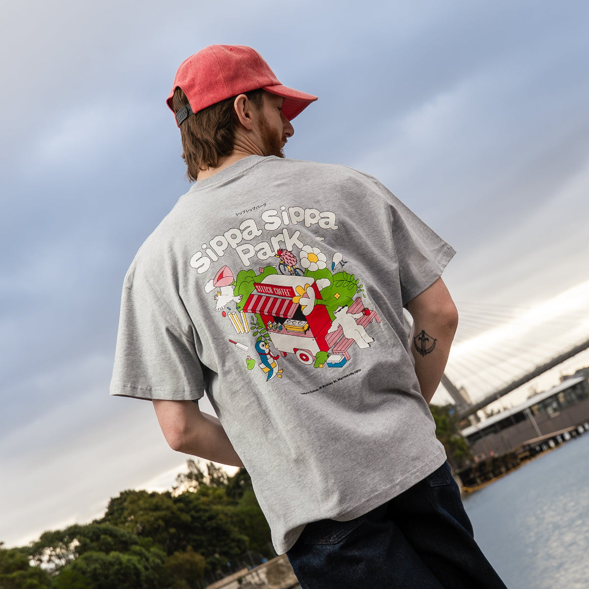 Person wearing a gray t-shirt with a colorful graphic and red cap, standing by a waterfront.