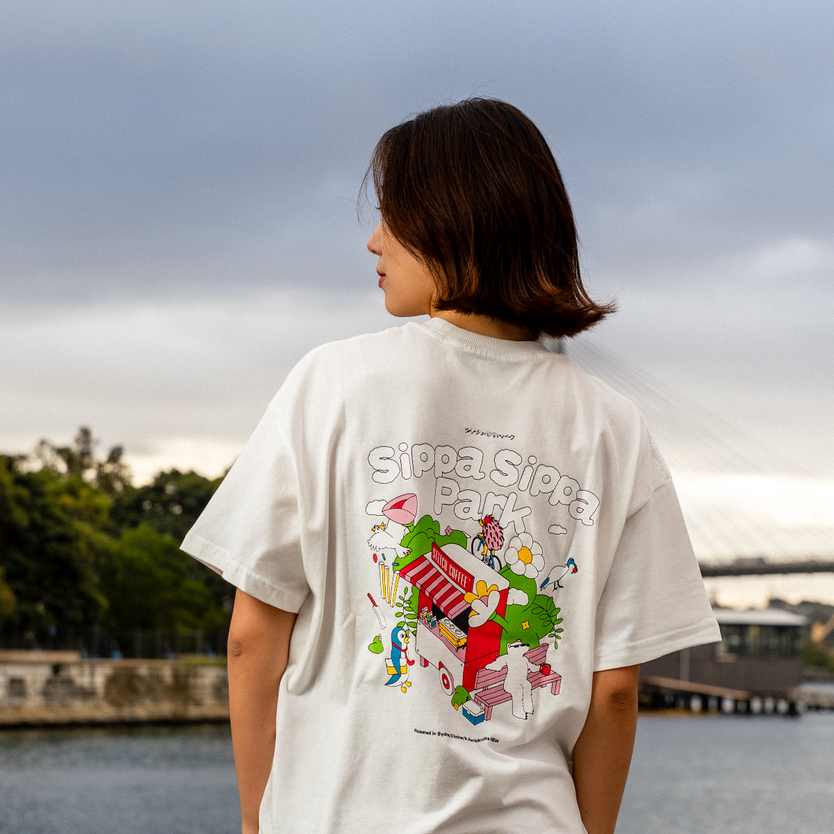 Person wearing a white t-shirt with colorful graphics and text, standing by a waterfront with trees and buildings in the background.